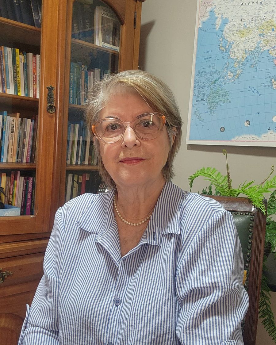 Mani Alvarez, de camisa listrada e óculos, sentada diante de uma estante de livros.